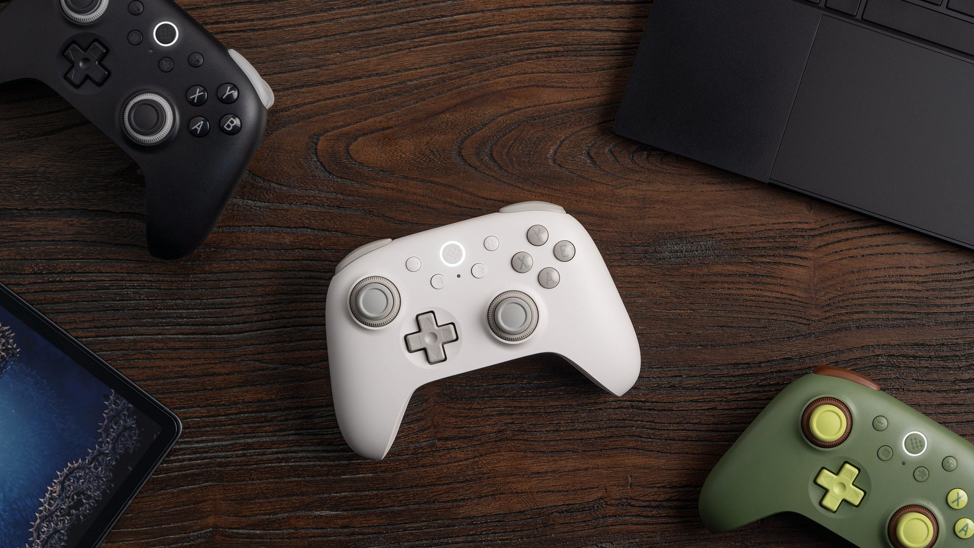 White, black and green 8BitDo Ultimate 3C+ controllers on a desk beside a tablet